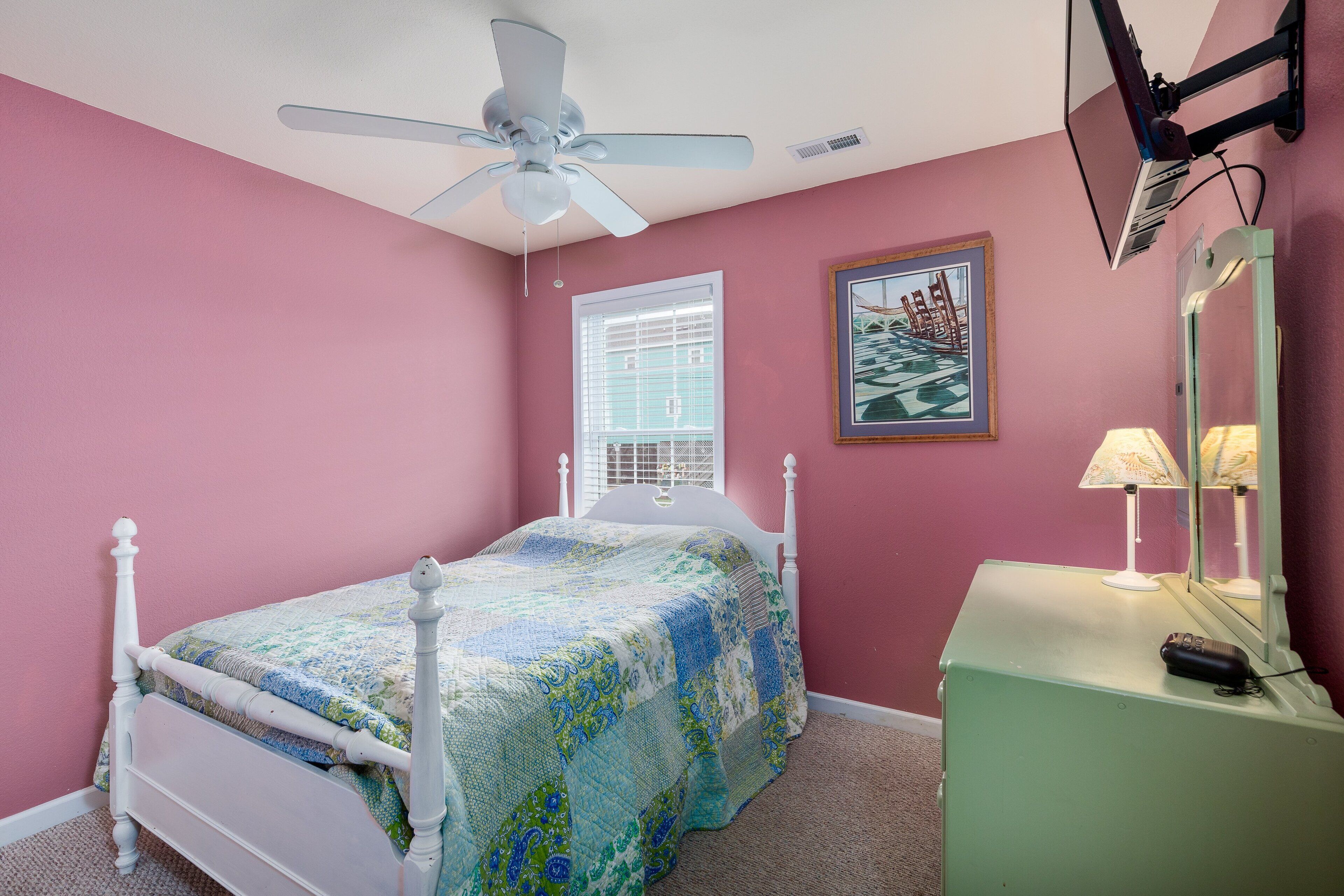 A bedroom with a bed, a ceiling fan, a window with blinds, a bedside lamp, and a mirror on a dresser.