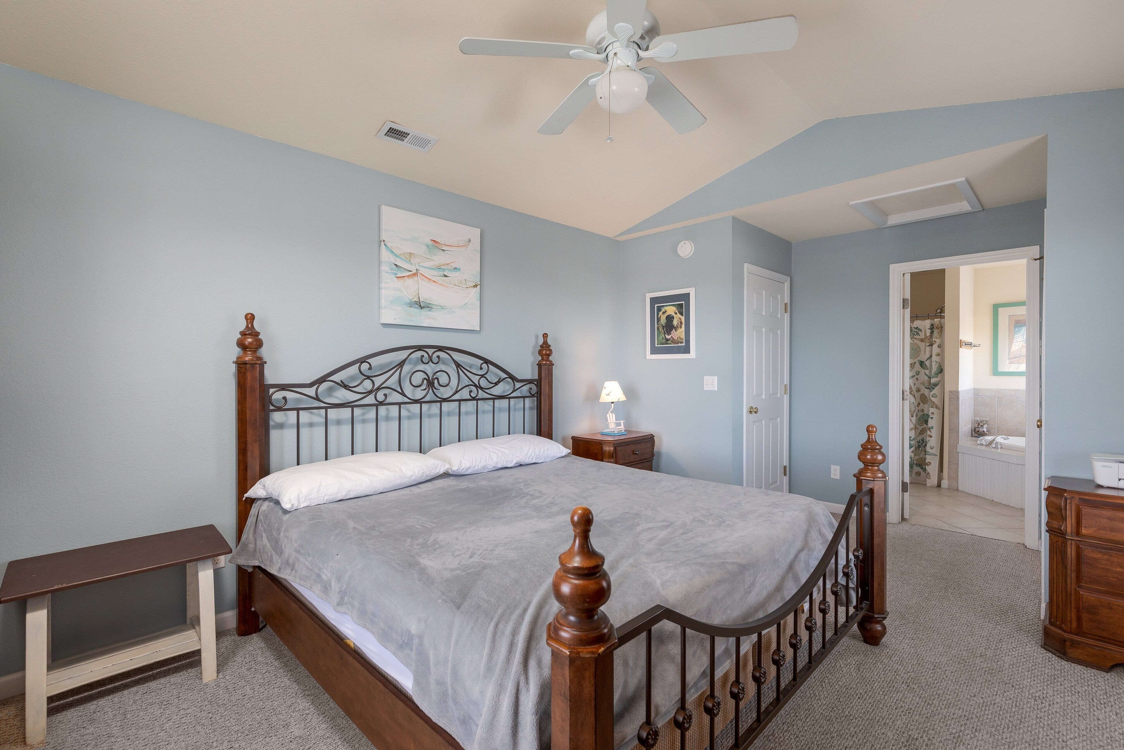 A bedroom with a bed, a nightstand, a bench, a ceiling fan, and a door leading to another room.