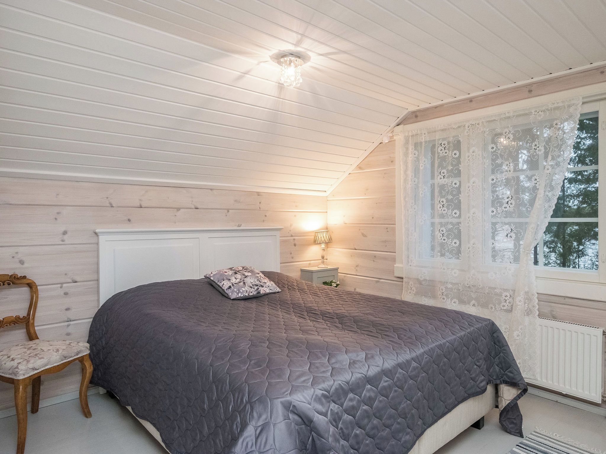 Een slaapkamer met een bed, een stoel, een nachtkastje en een raam met kantgordijnen.
