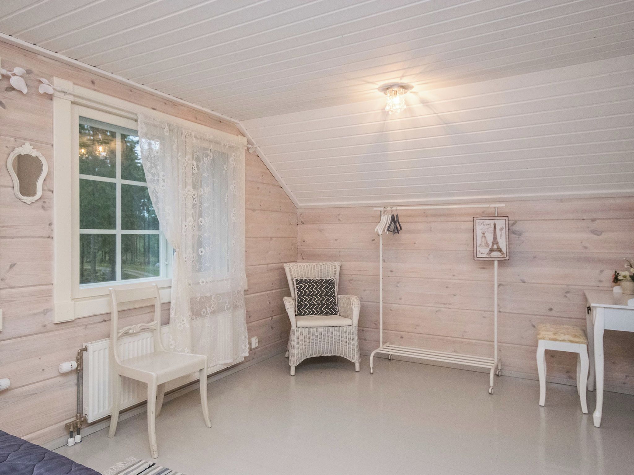 Una stanza con soffitto spiovente, una finestra con tende in pizzo, una sedia in vimini con un cuscino decorato, un termosifone e un tavolino con una cornice.