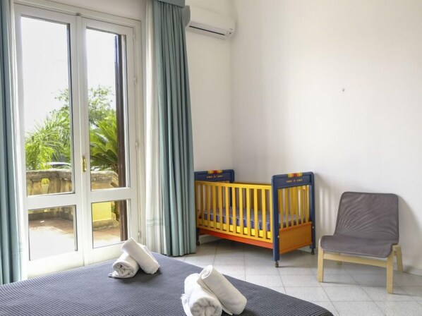 A room with a bed, a crib, a chair, and a large window with curtains.