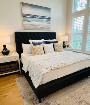 A bedroom with a large bed, a headboard, two bedside tables, a nightstand, a window, and a painting on the wall.