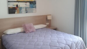 A bedroom with a large bed, a headboard, a bedside table with a lamp, a painting on the wall, and a window with curtains. - FUERTEVENTURA PORT SEA (Morro Jable)
