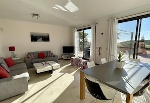 Una sala moderna con sofá, televisor, mesa de comedor y vistas amplias por ventanas grandes.