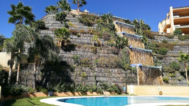 Ein Pool mit geschwungener Kante, eine bewachsene Hügelterrasse und ein Gebäude mit Balkonen.