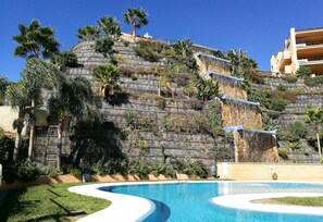 Una piscina de borde curvo, una colina con vegetación y un edificio con balcones.