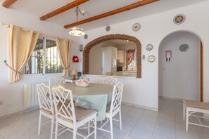 A dining area with a table and chairs, a window with curtains, a kitchen visible through an archway, and decorative plates on the wall.