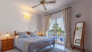 Quarto com cama, mesas de cabeceira, ventoinha no teto, janela com cortinas e vista para área externa.