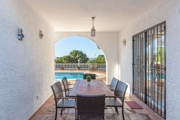 A patio with a table and chairs, a view of a swimming pool, and a white wall with decorative elements.