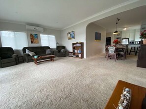 A spacious living room with a large sofa, a coffee table, and a bookshelf. There is a kitchen area visible in the background.