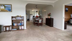Un salón amplio con piso alfombrado, una zona de comedor con mesa y sillas, una estantería y una cocina al fondo.