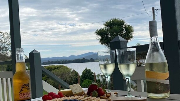 Una mesa con una tabla de quesos, frutas y copas de vino, con vista a un lago y montañas al fondo.