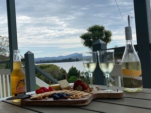 Uma mesa com tábua de queijos, frutas e copos de vinho, com vista para um lago e montanhas ao fundo.
