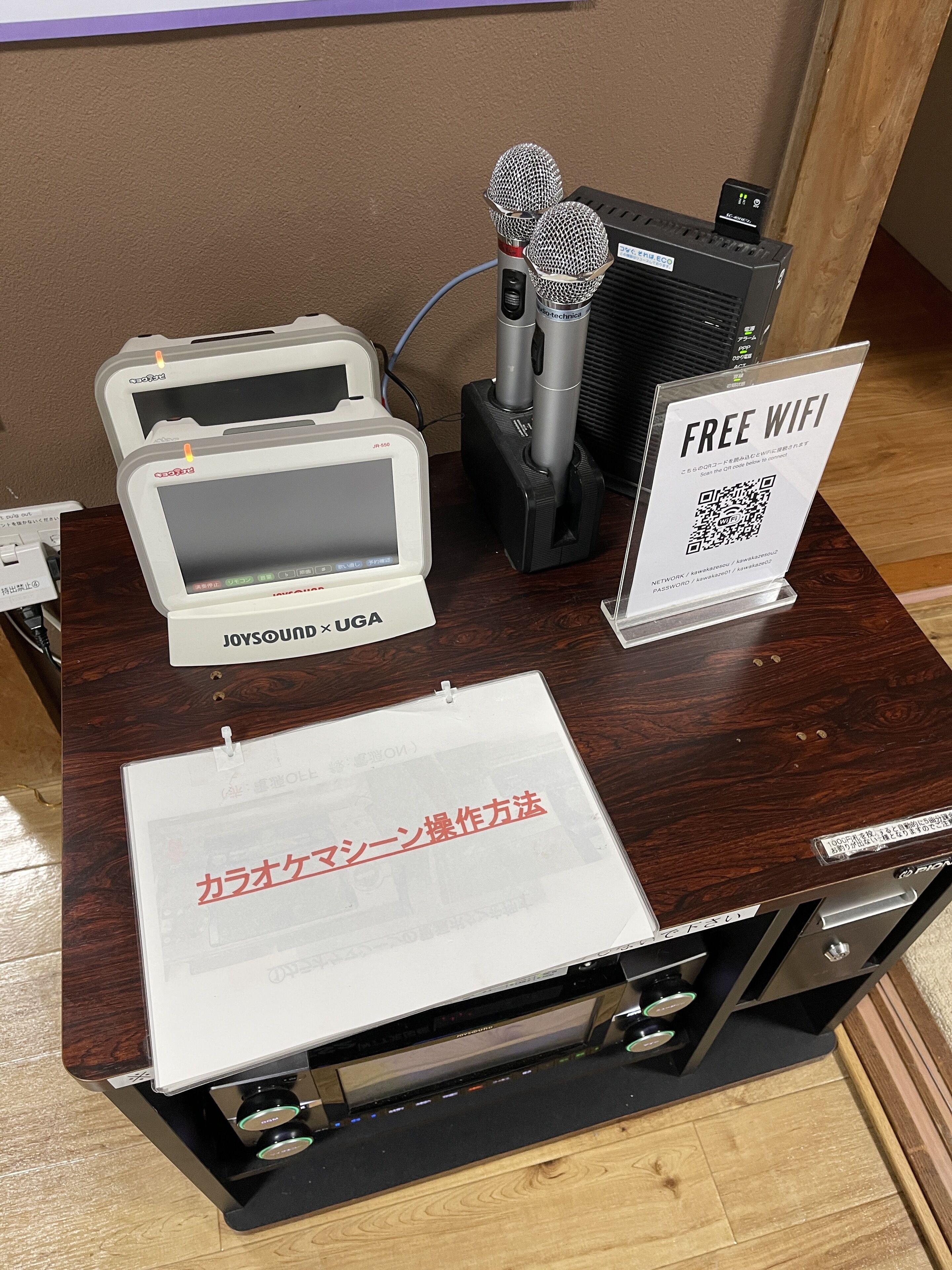 Ein Holztisch mit einem weißen Gerät mit der Aufschrift 'JOYSONIC UGA', einem Mikrofon und einem Schild mit 'FREE WIFI' und einem QR-Code.