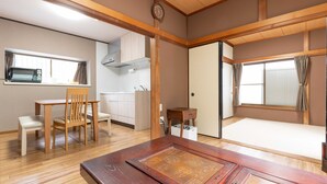 Une pièce de style japonais traditionnel, avec un sol recouvert de tatamis, une table à manger en bois entourée de chaises, un meuble bas en bois, un petit bureau en bois et une porte coulissante donnant sur un espace nuit.