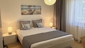 Une chambre à coucher avec un lit, deux tables de chevet, un luminaire suspendu, un tableau au mur et une fenêtre avec des rideaux.