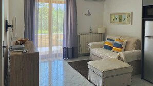 Une pièce bien éclairée, avec un canapé, un pouf et une petite table sur laquelle se trouve une lampe. Il y a une fenêtre avec des rideaux, un radiateur et un tableau encadré au mur.