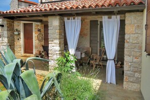 Ein Steinhaus mit Ziegeldach, weißen Vorhängen und einer Terrasse mit Stühlen und Tisch.