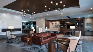 Un hall d’hôtel moderne avec un canapé en cuir marron, une table basse en bois et une cheminée. On y trouve plusieurs coins salon et, au fond, une cuisine.
