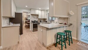 A modern kitchen with white cabinetry, a central island with stools, and a view of the outdoors through large windows. - Scenic Mountainside Home with Deck and Yard (Larkspur)