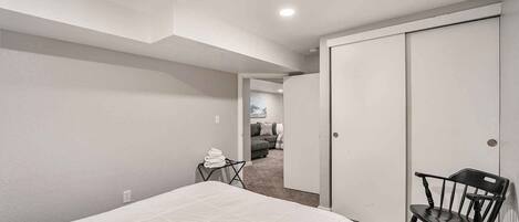 A bedroom with a bed, a chair, a closet, and a hallway leading to a living area.