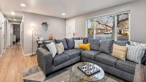A modern living room with a sectional sofa, a glass coffee table, and a large window providing a view of the street. - Modern Retreat | Yoga Rm | Vast Yard | Near Denver (Westminster)
