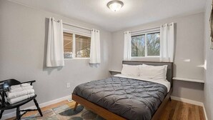 A bedroom with a bed, two windows with curtains, a bedside table, a chair with towels, and a patterned rug. - Modern Retreat | Yoga Rm | Vast Yard | Near Denver (Westminster)
