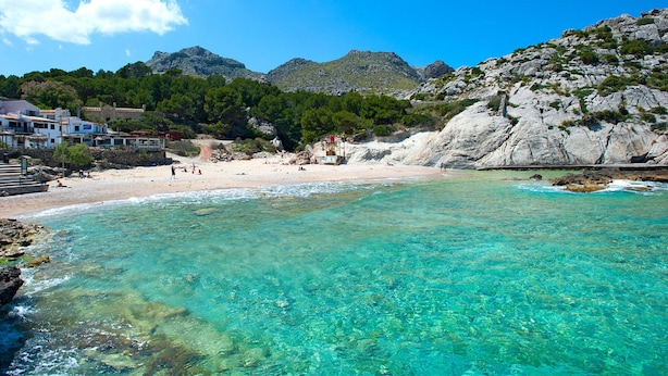 Una playa costera con aguas turquesas cristalinas, acantilados rocosos y montañas al fondo.
