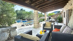 Un patio al aire libre con mobiliario de mimbre, una pared de piedra y vistas a una piscina y montañas.