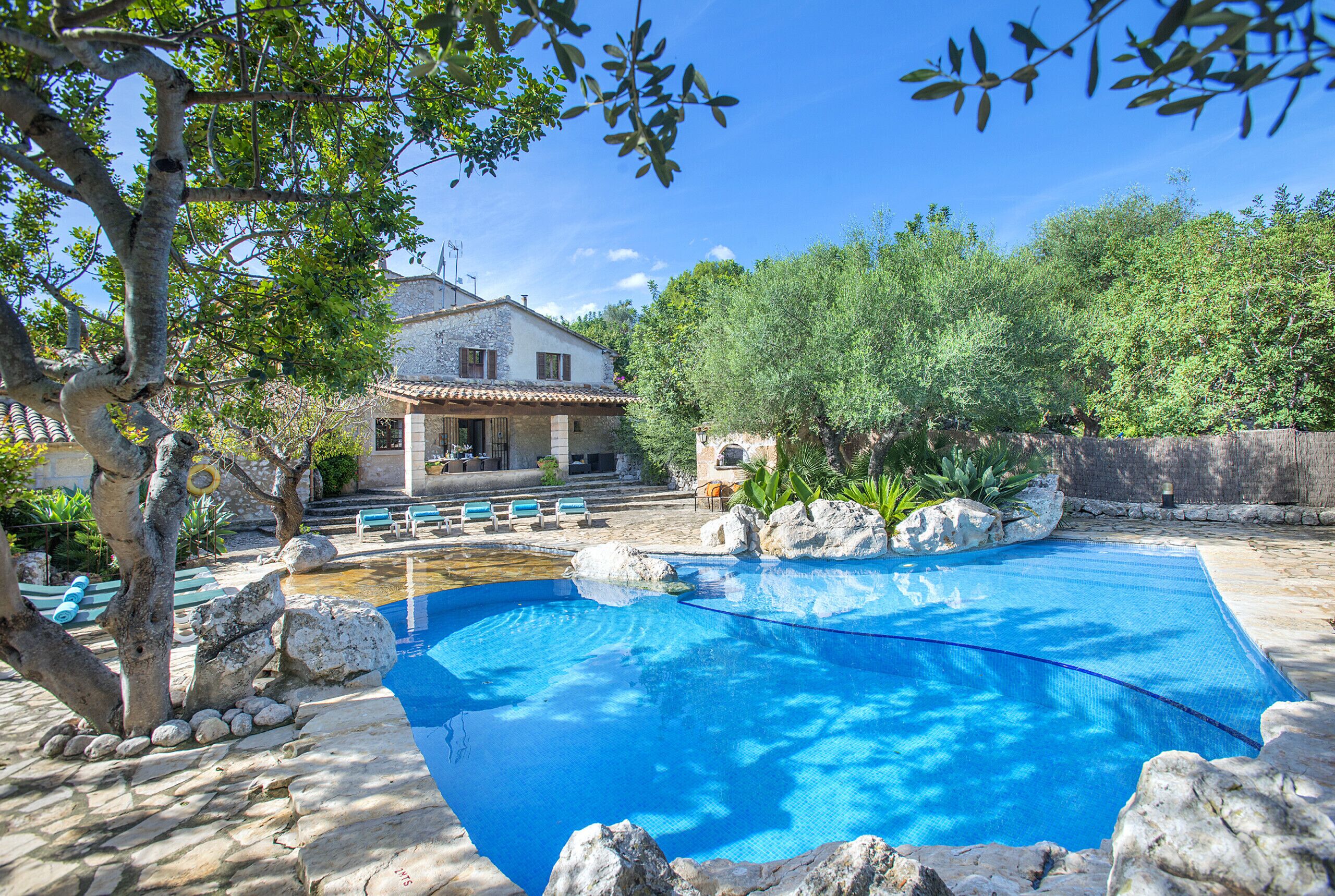 A swimming pool with a stone deck, surrounded by rocks and trees, with a house in the background.