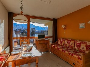 Um quarto aconchegante com uma mesa de jantar de madeira posta para quatro pessoas, um sofá com capa acolchoada e vista para montanhas através de grandes janelas.