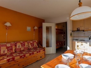 Quarto aconchegante com sofá de madeira, mesa de jantar posta para dois e uma cama beliche ao fundo.