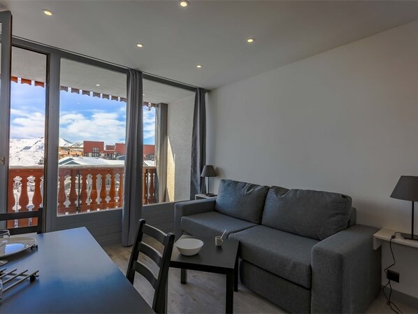 Un salon moderne avec un canapé gris, une table à manger avec des chaises et un balcon offrant une vue sur des bâtiments enneigés.