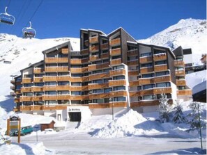 Un hôtel à plusieurs étages doté de balcons, avec un télésiège visible au loin et un paysage montagneux enneigé.