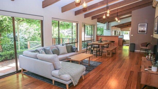 A modern living room with a sofa, coffee table, and dining area. Large windows offer views of greenery. - Haus on Houghs : Space and style in Bright (Bright)