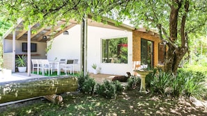 Un espace extérieur aménagé avec des sièges, protégé par un toit, comprenant des chaises blanches et une table, un bâtiment en briques doté d’une grande fenêtre, et un jardin avec un abreuvoir à oiseaux et une bûche.