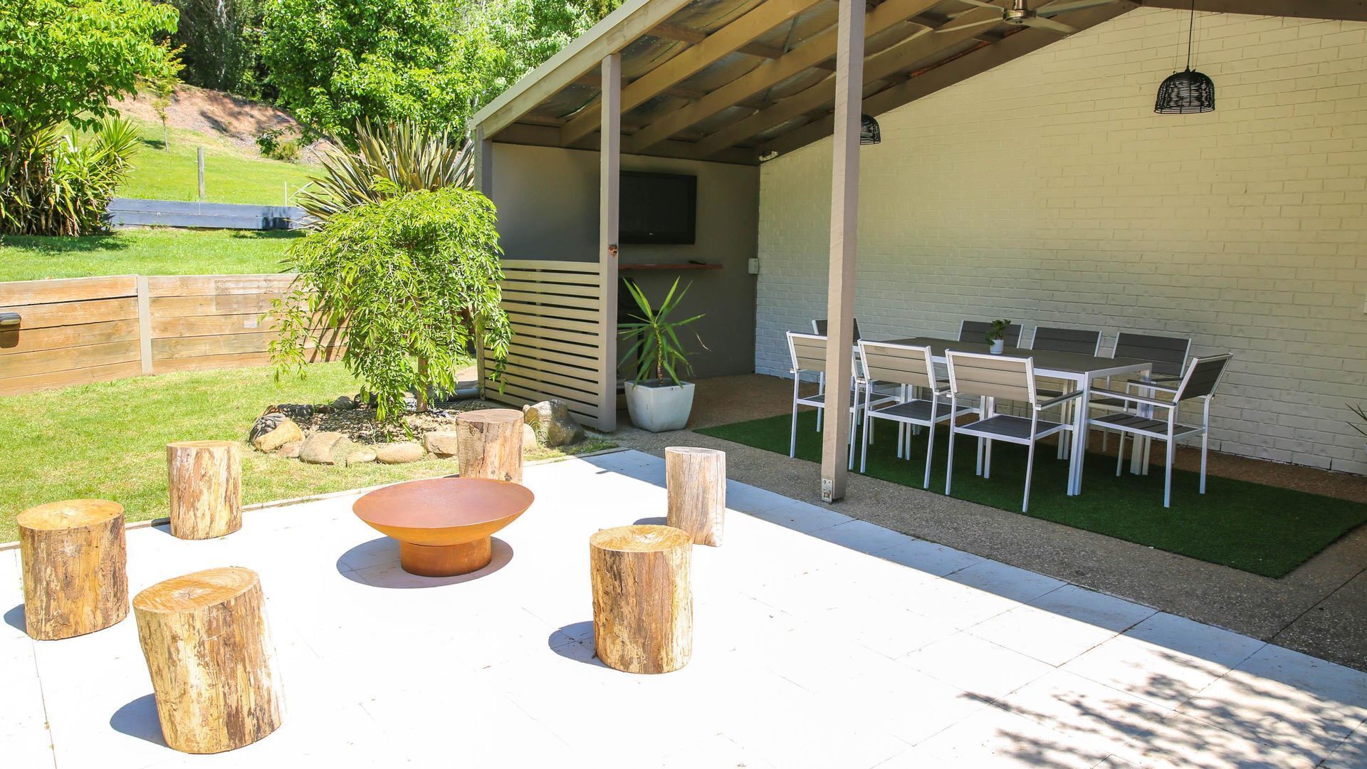 Une terrasse extérieure avec une table et des chaises, entourée de troncs d’arbres, d’une plante en pot et d’un téléviseur fixé au mur.