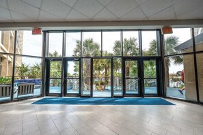 A hotel entrance with glass doors, a blue mat, and a view of palm trees and a pool.