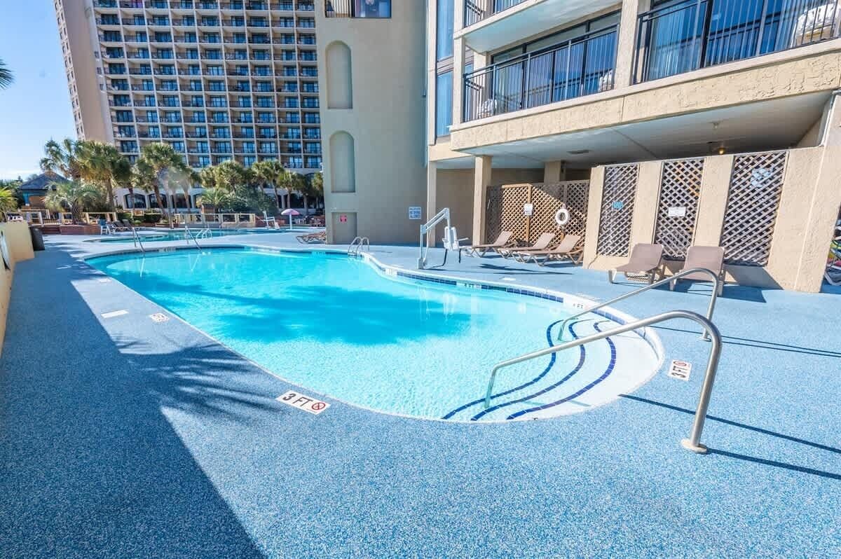 A swimming pool with a 3ft safety zone, lounge chairs, and a building with balconies in the background.