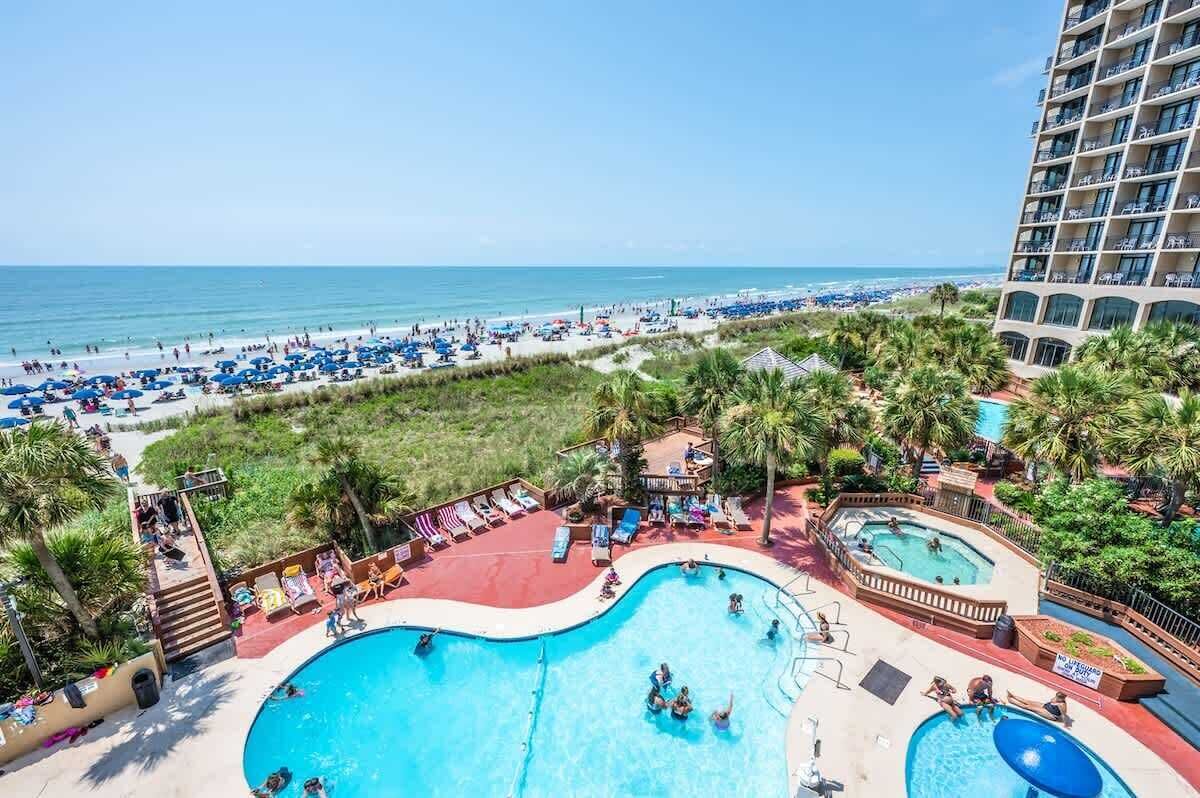 Een hotel met een groot zwembadgedeelte, uitzicht op een strand en een gebouw met meerdere balkons.