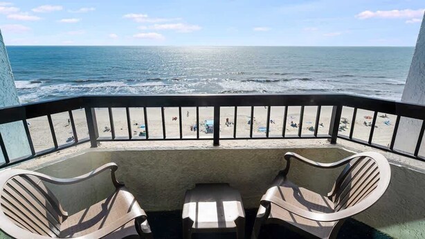 A balcony with two chairs and a table overlooking a beach with people and umbrellas. - Recently Renovated, Oceanfront View, Heated Pools! (Myrtle Beach)