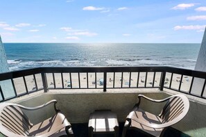 A balcony with two chairs and a table overlooking a beach with people and umbrellas. - Recently Renovated, Oceanfront View, Heated Pools! (Myrtle Beach)