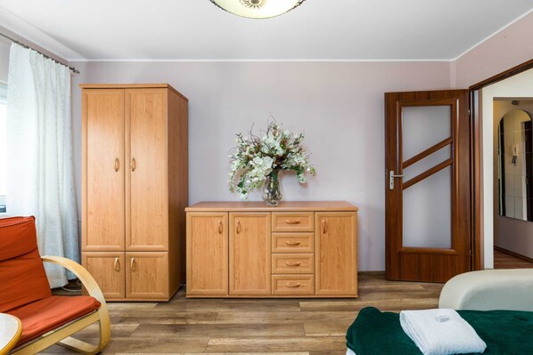 A room with a wooden wardrobe, a bed with a green blanket, a chair, and a vase with flowers on a cabinet. - Spacious Apartment | Bath, Close to Airport and Trade Fair (Poznan)