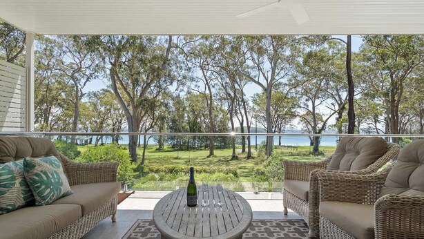 Un balcon agrémenté de meubles en osier, une table avec une bouteille et des verres, et une vue sur les arbres et l’eau.