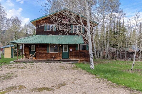 Une cabane en rondins à deux étages, avec un toit en métal vert, une véranda à l’avant et une allée gravillonnée.