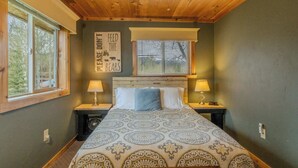 A bedroom with a wooden ceiling, a bed with patterned bedding, two bedside lamps, a window with blinds, and a wall sign. - Alpine Star Lodge (West Yellowstone)