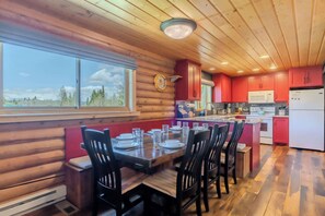 A log cabin interior with a dining area, kitchen, and large windows overlooking a forested landscape. - Blue Spruce Cabin (West Yellowstone)
