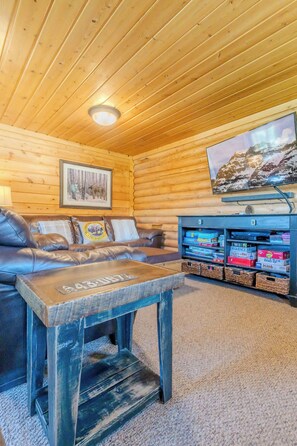 L’intérieur chaleureux d’un chalet avec un plafond en bois, un canapé en cuir, une table basse, un téléviseur fixé au mur et une bibliothèque.