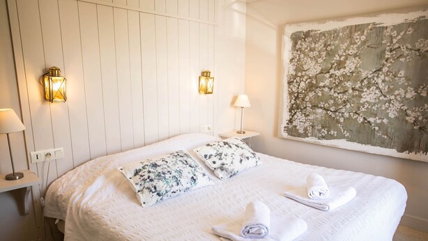 Ein ordentlich bezogenes Bett mit floralen Kissen, zwei Wandlampen und ein großes Blumenbild an der Wand.