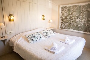 Un lit bien fait, agrémenté de coussins à motifs floraux, deux lampes fixées au mur et un grand tableau floral accroché au mur.
